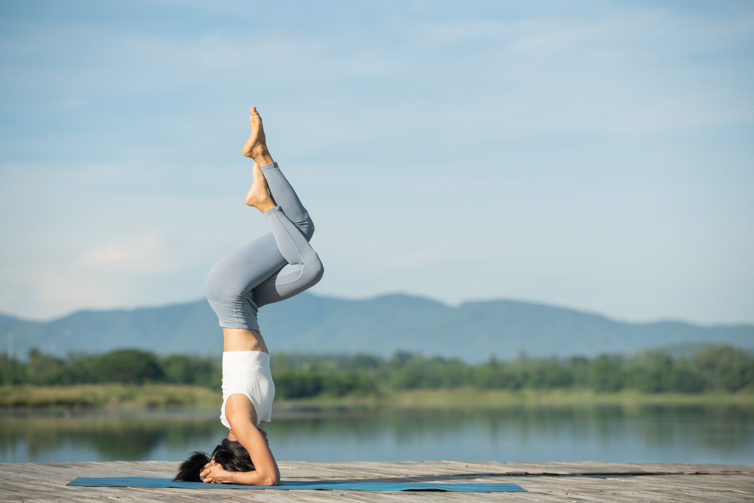 "A woman practicing yoga outdoors, performing a headstand pose on a wooden deck near a serene lake with mountains and a clear blue sky in the background. Keyword: Yoga Journey